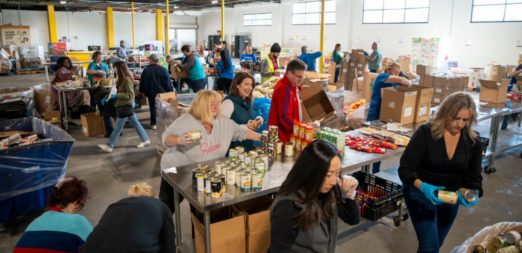 A group of volunteers sorts and packs food items at large tables in a busy warehouse, preparing boxes of canned goods and snacks for distribution.